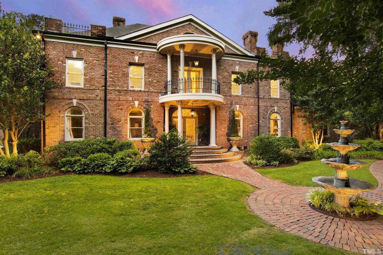 A large brick home with a grand front entrance featuring a fountain, stone pathway, manicured landscaping, and many trees